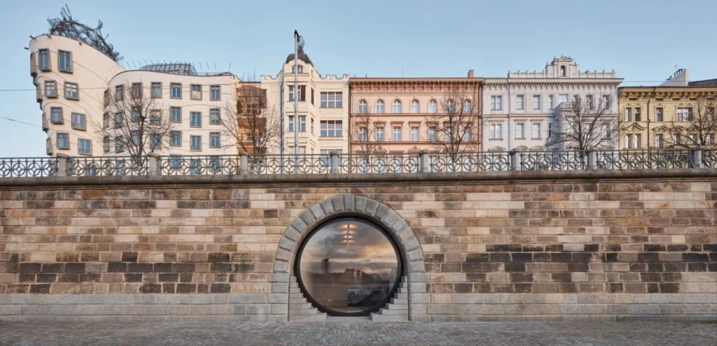 Ușă circulară din sticlă integrată în zidul cheiului râului Vltava, cu clădiri istorice din Praga în fundal.